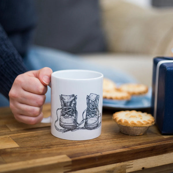 A woman's hand holding a mug with a hand-drawn illustration of old leather walking boots on a coffee table with mince pies - perfect gift for hikers.
