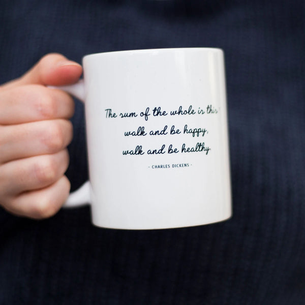 Woman's hand holding a Charles Dickens be happy walking quote mug.