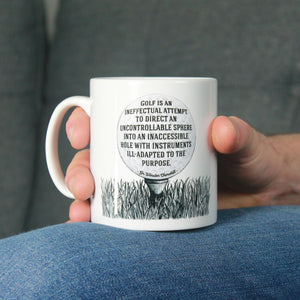 Male hand holds a black and white mug printed with an illustration of a golf ball on a golf tee which contains a funny golf quote from Sir Winston Churchill which reads: Golf is an ineffectual attempt to direct an uncontrollable sphere into an inaccessible hole with instruments ill-adapted to the purpose. The mug is resting on a denim-clad leg against a grey sofa in the background.
