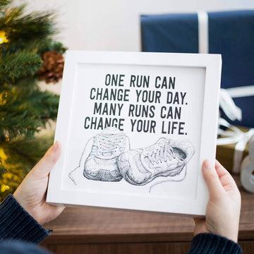 Woman holding framed running art christmas gift for a runner in front of wrapped christmas gifts and a tree 