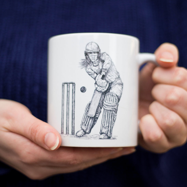 closeup of woman's hands holding a white mug with an ink illustration of a female cricketer batting on it
