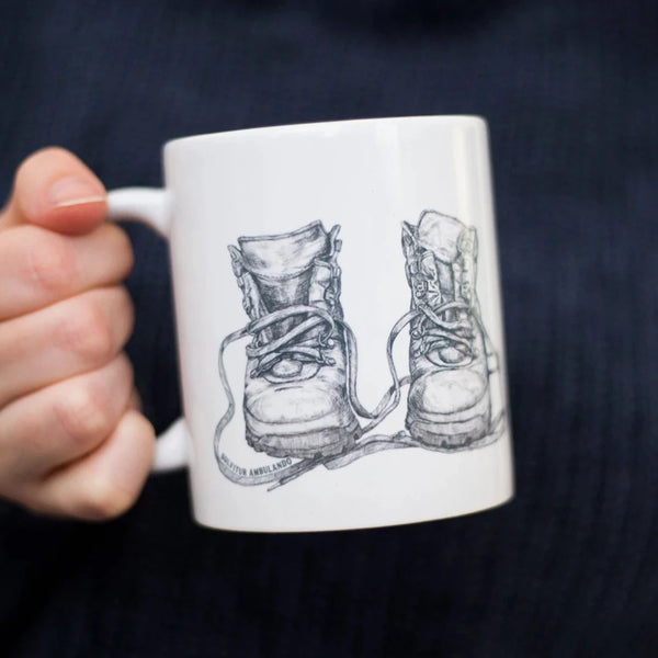 Closeup of a woman's hand holding a Solvitur Ambulando Old Leather Walking Boots Mug - a thoughtful gift for those who love hiking.