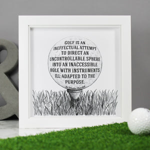 Sir Winston Churchill funny golf quote:  Golf is an ineffectual attempt to direct and uncontrollable sphere into an inaccessible hole with instruments ill-adapted to the purpose. Framed illustrated art print gift for golfers. Made by Wit and Wisdom UK. Shown here in a quare white box frame against a grey backdrop with large grey display ampersand in the background, and a golf ball on a section of green astro turf in the foreground of the photo.