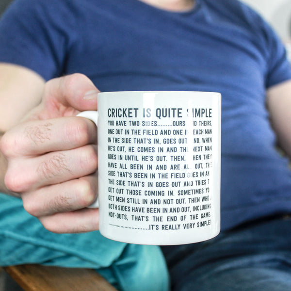 Male hand holding a black and white mug with the funny cricket quote 'Cricket is Quite Simple' printed on it. The man is wearing a blue tshirt and jeans and resting his forearm on a turquoise cushion.