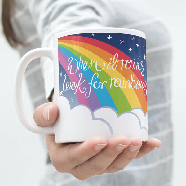 Woman in a grey and white stripy top holding the 'When it rains, look for rainbows' illustrated rainbow mug by Wit and Wisdom in her hand which is outstretched towards the camera.