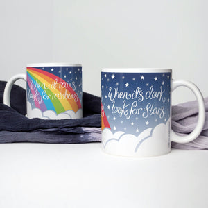 Two mugs on a white background with indigo scarf, mugs show the wrap-around design of a dark blue starry night sky with clouds and a rainbow, with the words: When it rains, look for rainbows. When it's dark, look for stars - designed by Wit and Wisdom UK.